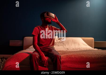 Black man using virtual reality headset at home with neon light, sit on bed at night. VR addiction. Stock Photo