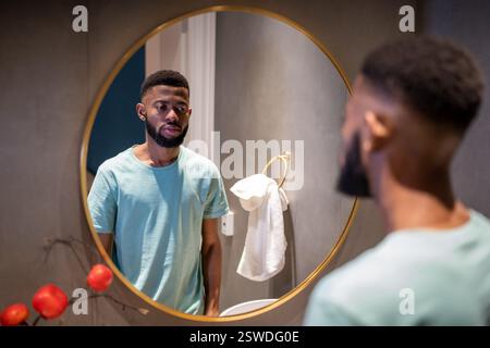 Unhappy African American man looking in mirror in bathroom, feeling dissatisfied with his appearance Stock Photo