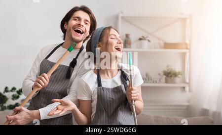 Cheerful Couple Using Mop Handles As Microphones, Having Fun During Spring-Cleaning Stock Photo