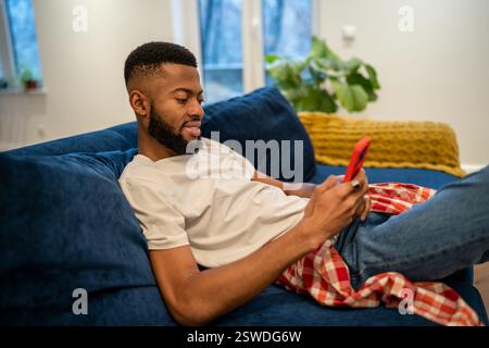 Relaxed smiling african american smiling man sits on couch looks at smartphone at home. Stock Photo