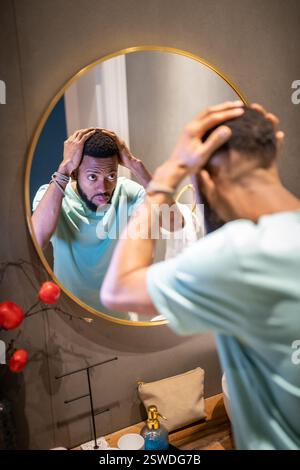 Upset young African American man looking in bathroom mirror touching hair worrying about balding Stock Photo