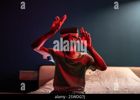 Black guy wearing virtual reality headset sitting on bed at home exploring metaverse. VR concept Stock Photo