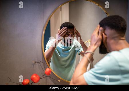 Unhappy African American 30s man touching face skin looking at mirror worrying about first wrinkles Stock Photo
