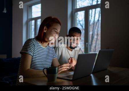 Smiling multiracial family couple enjoy freelancing, sitting at table working on laptops together Stock Photo