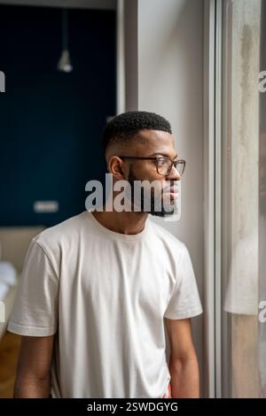 Upset young African American man standing at home looking out window and pondering Stock Photo