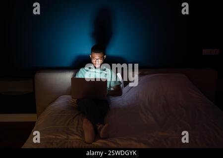 Young black man works on laptop lying in bed at night in dark looks attentively at screen. Stock Photo