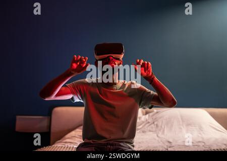 Young African American man sitting on bed under red neon light using futuristic metaverse VR headset Stock Photo