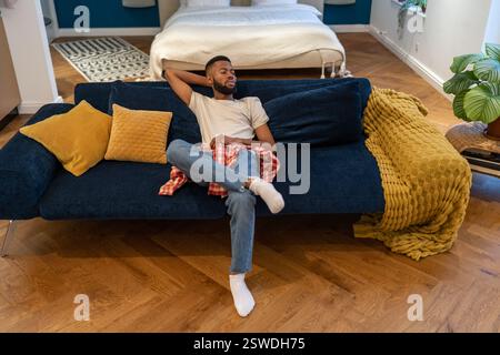 Thoughtful dreaming african american guy sits on sofa rests in studio apartment. Stock Photo