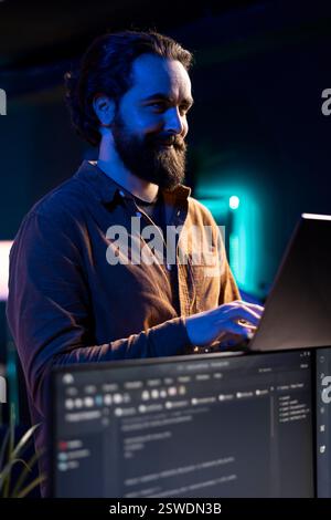 Joyful freelancer doing software debugging using laptop, checking errors in lines of code displayed on PC monitors. Cheerful IT professional walking in apartment fixing coding issues using notebook Stock Photo