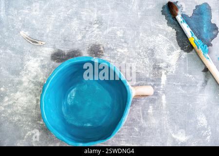 A rustic, hand-painted ceramic cup with a vibrant blue interior sits on a textured gray surface. Paintbrush with blue paint lies beside it Stock Photo