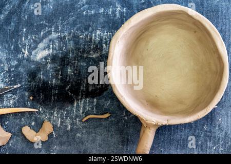 Handcrafted, unfired clay bowl with a handle sits on a textured blue surface. Surrounding the bowl are small pieces of clay and sculpting tools Stock Photo