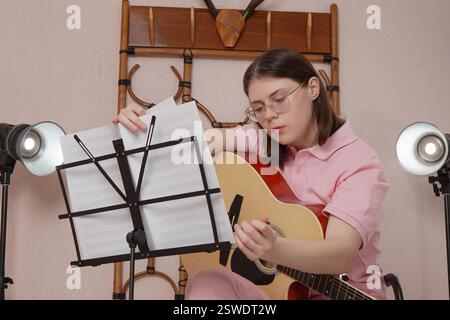 Young woman in glasses with guitar flips through sheet music on music stand before starting to play Spanish string acoustic guitar in home studio Stock Photo