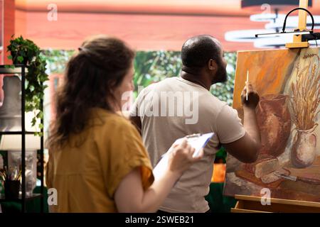 Woman master painter teaching her apprentice to use new painting techniques in art studio, sharing her skill and ideas in creating a rustic artwork on canvas. Artist helping man with paintwork. Stock Photo