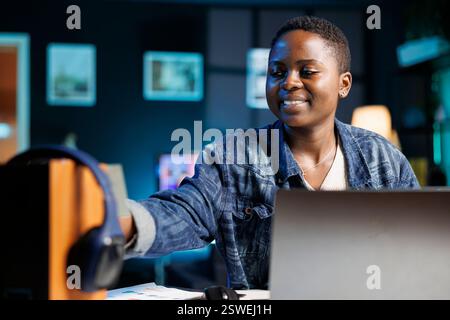 Young african american freelancer works from home on laptop while sticking a sticky note to electronic speaker. While using her personal computer, smiling black woman puts a post-it note on device. Stock Photo