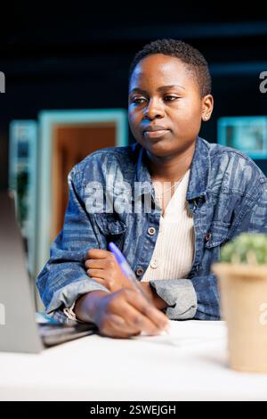 Focused African American lady at home using a laptop and taking notes. Portrait of black woman with notepad, studying online, researching, and multitasking to work from her apartment. Stock Photo