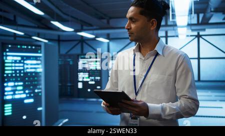 Programmer walking in high tech data center used for managing infrastructure energy consumption, working on tablet. Indian man using device in workspace used for automation in server farm, camera B Stock Photo