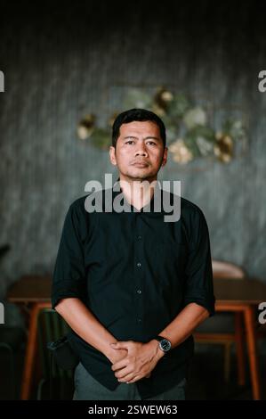A portrait of an Indonesian man in a dark shirt, posing indoors. He exudes a thoughtful and serious demeanor. The background offers a soft contrast. H Stock Photo