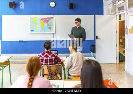 Geography teacher explaining a lesson holding a clipboard and smiling to elementary school students in a classroom Stock Photo
