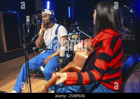 Two musicians collaborating in a professional recording studio, one strumming an acoustic guitar while the other sings into a microphone Stock Photo