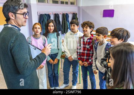 Male teacher holding clipboard explaining lesson to group of elementary school students in classroom Stock Photo