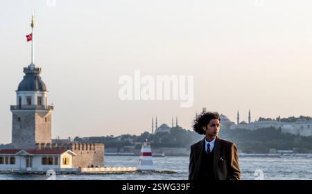 A young man in a suit stands by the water in Istanbul, with the Maiden's Tower and historical skyline in the background during sunset. Stock Photo