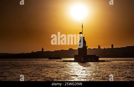 A silhouette of a lighthouse at sunset, with the sun setting behind it. The water reflects the warm golden hues of the sky, and the skyline of a city Stock Photo