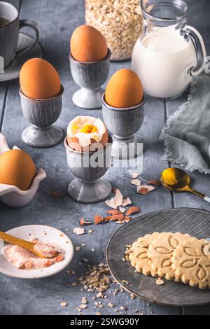Boiled brown eggs in gray egg cups and other breakfast ingredients on gray background, preparation for breakfast, vertical Stock Photo