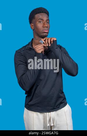 Studio portrait of a young adult male model wearing a black sweatshirt and white pants, adjusting his pendant necklace against a vibrant blue backdrop Stock Photo