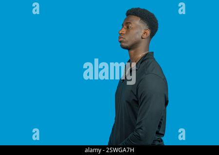 Studio shot of a young black man wearing black long-sleeved shirt, posing in profile against a vibrant blue backdrop Stock Photo