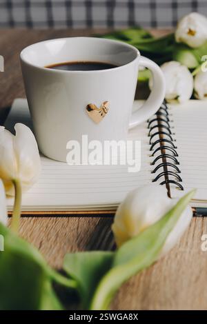 Cup of Coffee, open book and a bouquet tulip flowers over wood table top. Composition with copy space. Minimal flower arrangement. Stock Photo