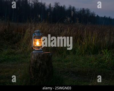An old oil lantern with a lit fire stands on the stump outdoors at night. Soft warm yellow light of an old kerosene lamp. Stock Photo