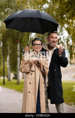 A loving couple enjoys snacks while standing under an umbrella on a rainy day in the park. Stock Photo