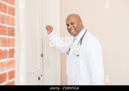 Smiling doctor in a white coat knocking on a door for a house call Stock Photo