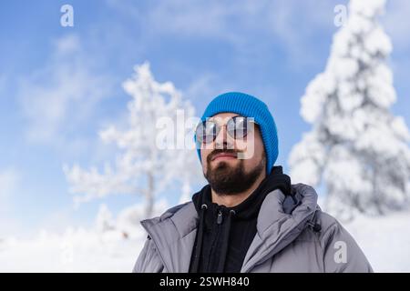 Bearded man in sunglasses & hat against blue sky and snowy trees Stock Photo