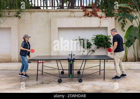 Two people engage in a lively game of table tennis in the courtyard. Middle aged woman and young man sport battle. The atmosphere is relaxed, and they Stock Photo