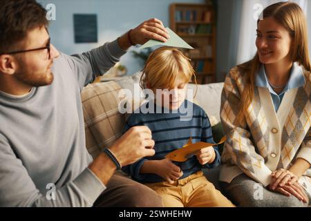 Family sitting on couch and creating paper crafts together, father holding paper above sons head while mother smiling and guiding son in craft making Stock Photo