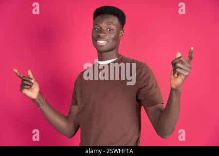 A cheerful young African man stands against a vibrant pink backdrop, pointing upwards with both hands. His bright smile and casual brown T-shirt sugge Stock Photo