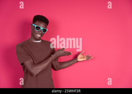 Young smiling black man wearing blue glasses with carmin red background pointing at the copyspace to put any text or virtual button isolated on red ba Stock Photo