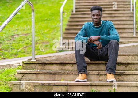 A young man in a blue shirt sits on a set of stairs. He is wearing brown shoes and jeans Stock Photo