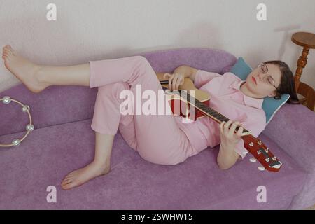 Woman guitarist with eyeglasses and her eyes closed laying on her back on sofa and playing guitar. Musician of Caucasian ethnicity dressed casually in Stock Photo