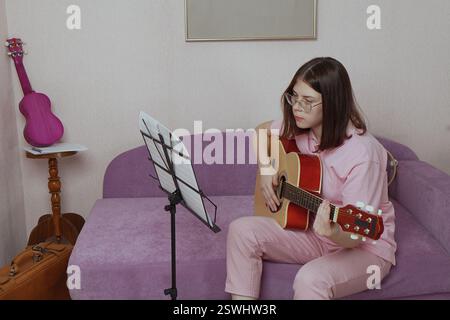 Woman is strumming her acoustic guitar in living room, her fingers dancing across strings as she performs melodic composition. She's sitting at sofa i Stock Photo