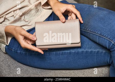 Beige wallet held by woman sitting on sofa in casual clothes, emphasizing fashion accessory concept Stock Photo