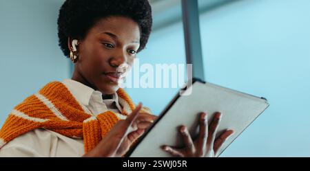 A confident black businesswoman is focused while using a tablet. She is dressed in professional attire with wireless earbuds, conveying a sense of mod Stock Photo