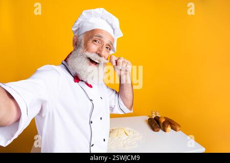 Senior chef joyfully posing in professional uniform at bakery counter with yellow background and baking essentials Stock Photo