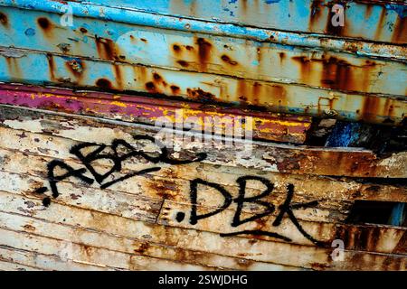Black spray can graffiti on the peeling but colourful and richly textured paintwork on the rotting hull of an old fishing boat abandoned on Le Sillon, the causeway leading to Le Fret, a former sardine, scallop and oyster fishing village on the Crozon peninsula in Finistère, Brittany, France. Stock Photo
