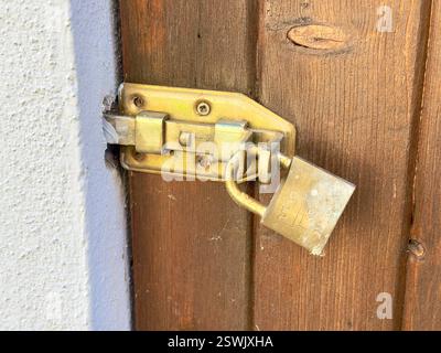 Close-up of an old golden latch secured with a padlock on a wooden door. The door appears freshly painted, contrasting with the aged and slightly dirt Stock Photo