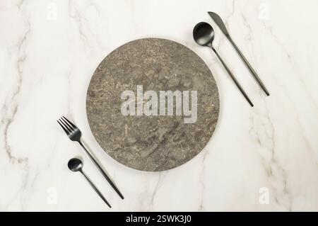Top view of brown marble plate, black cutlery on white marble background. Food presentation, stone tray. Table setting, copy space, flat lay. Stock Photo