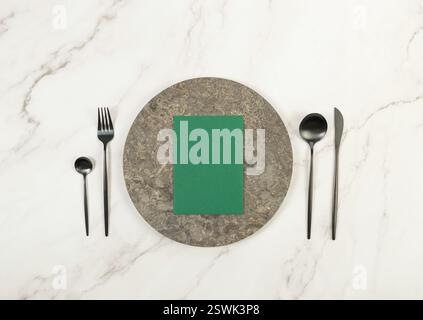 Top view of brown marble plate, black cutlery and green card on white marble background. Food presentation, stone tray. Table setting, copy space. Stock Photo