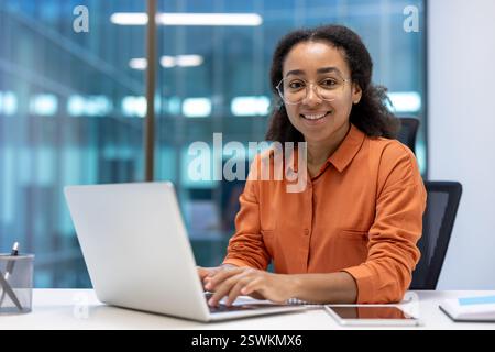 Portrait of a successful businesswoman at workplace inside office. The woman smiles and looks at camera typing on the laptop keyboard. The employee in a casual shirt works sitting near the window. Stock Photo