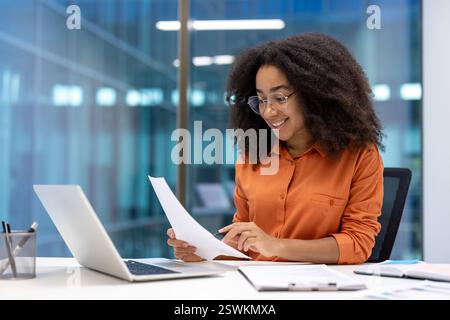 Successful businesswoman at workplace inside office working with documents, reports and contracts. Woman smiling happily, using laptop at work. Stock Photo
